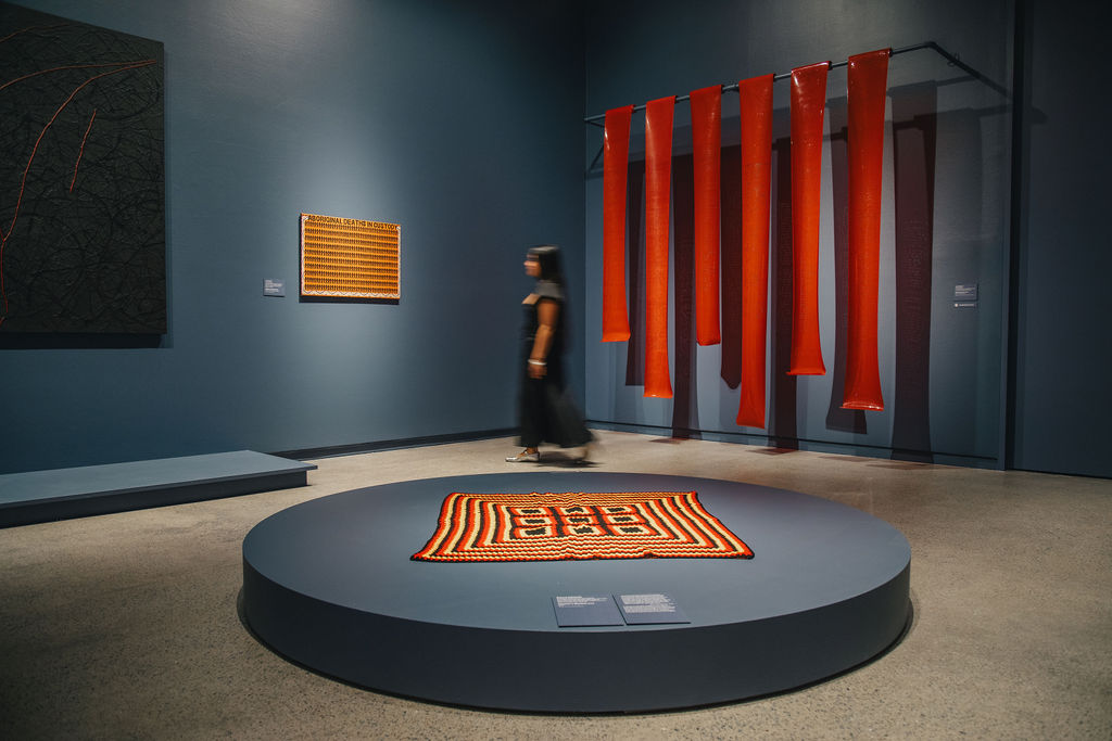A woman walks through an art gallery featuring red hanging sculptures and a woven rug on a circular platform.